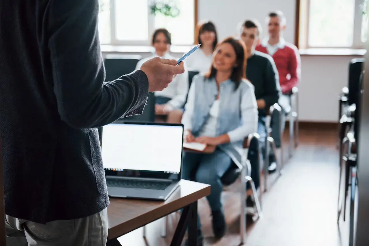 Professor de costas apresentando aula para turma de adultos sentados em uma sala de treinamento