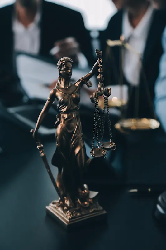 Estátua da Justiça sobre a mesa de um escritório de advocacia, com advogados revisando documentos ao fundo